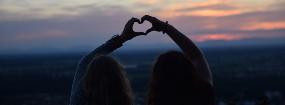 Kuvassa on taustalla epäselvänä näkyvä tunturimaisema auringonlaskun aikaan. Etualalla näkyy kaksi naista, jotka tekevät käsilään sydämen.